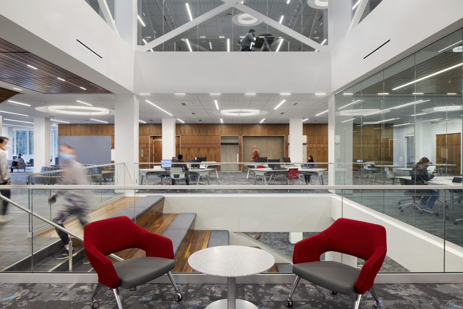 Two red seats in large atrium area with stairs down to lower level, white walls, wood paneling, and grey carpet Two red seats in large atrium area with stairs down to lower level, white walls, wood paneling, and grey carpet