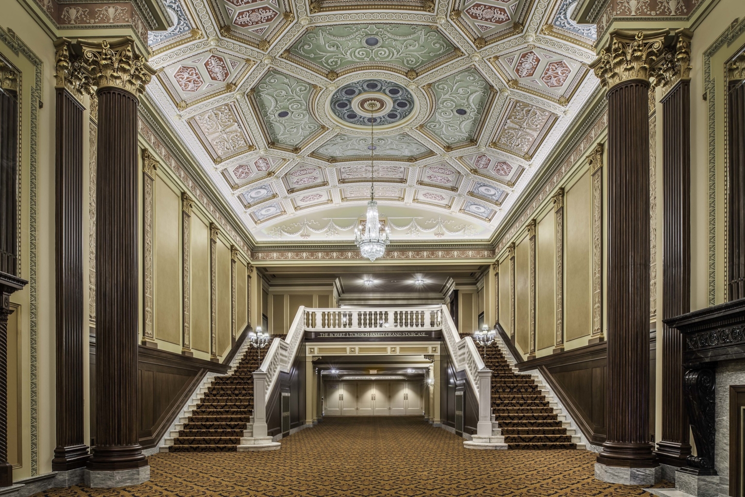 a carpeted lobby with colorful frescoes in a coffered ceiling and a double sided grand stair