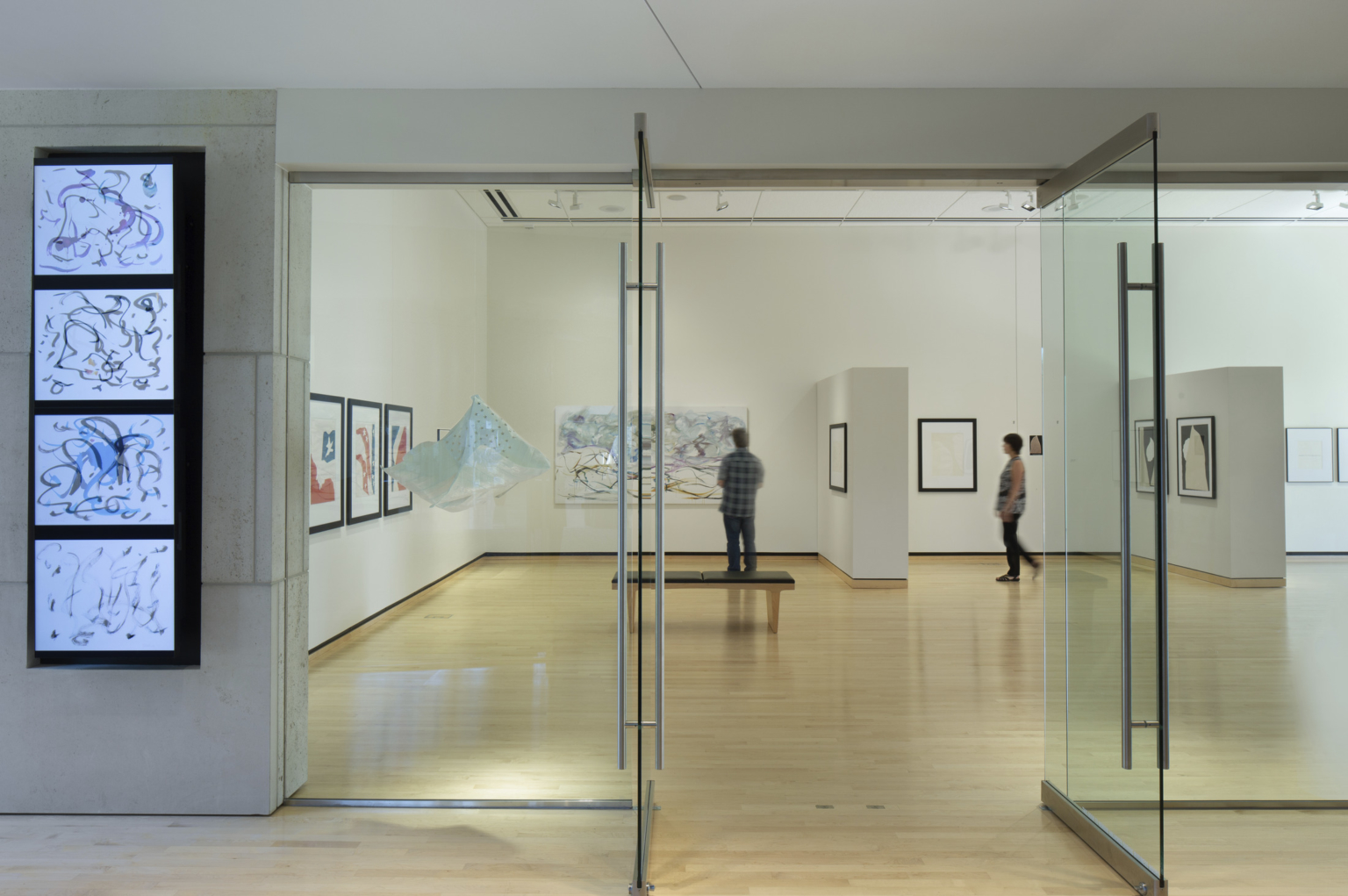 Looking into an art gallery through glass doors with interactive screen on the lefthand side, warm wood floors, and white walls at the Washburn University Rita Blitt Gallery Addition Looking into an art gallery through glass doors with interactive screen on the lefthand side, warm wood floors, and white walls at the Washburn University Rita Blitt Gallery Addition