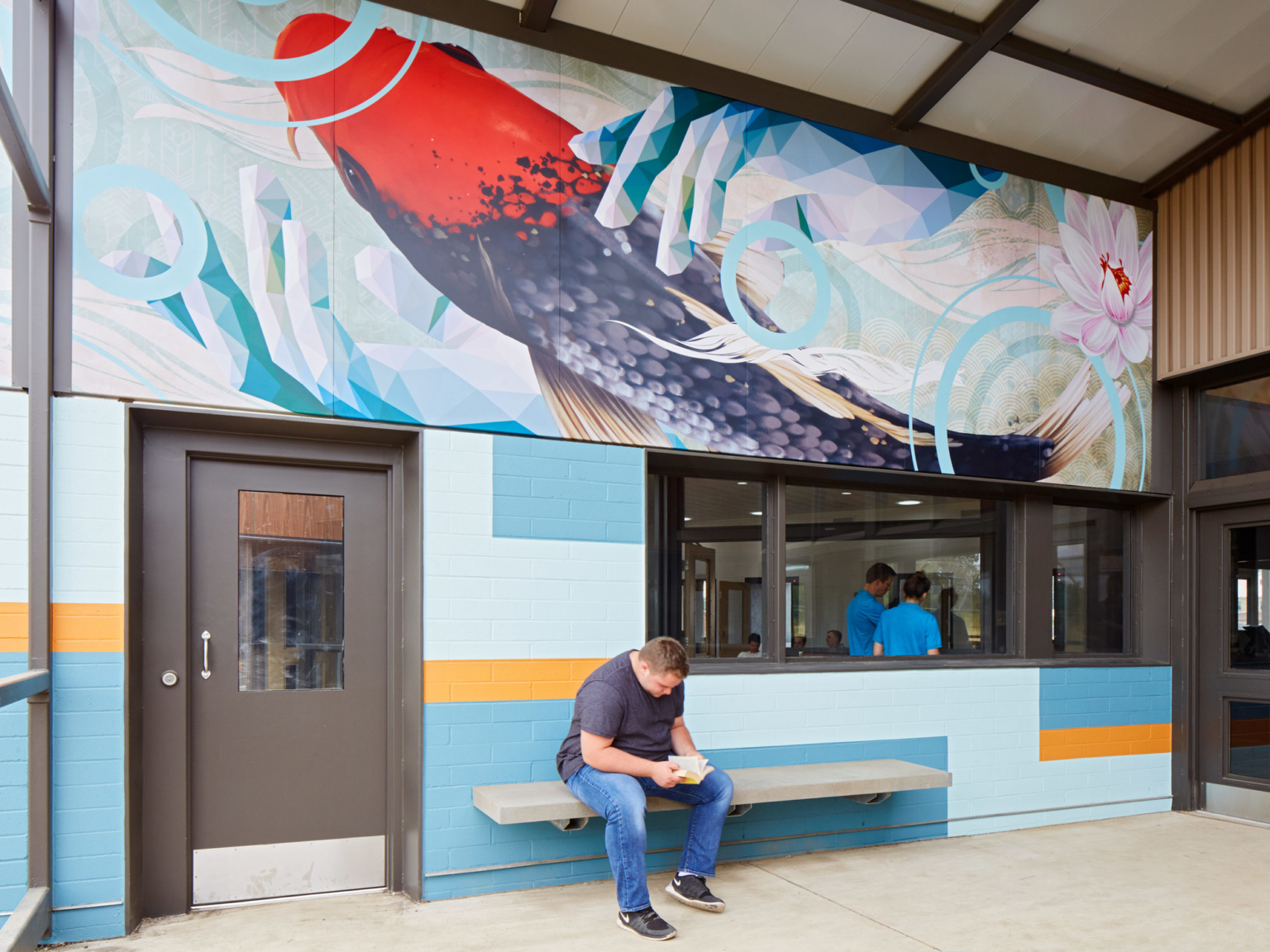 a man is seated on a bench against a blue wall with a large mural of a colorful fish, geometric hands, and a flower