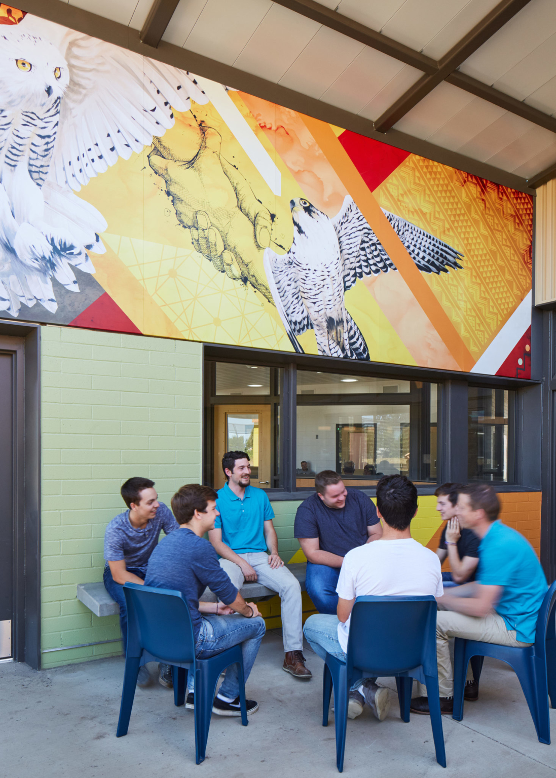 a group of young men seated in chairs in a circle next to a bright orange wall with a mural of hands and birds of prey