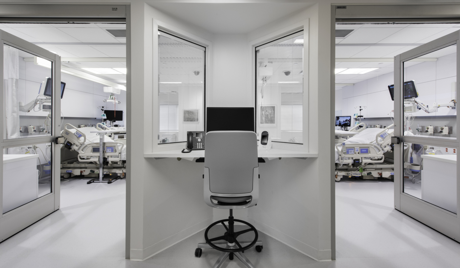 a desk between observation windows looking into two patient rooms