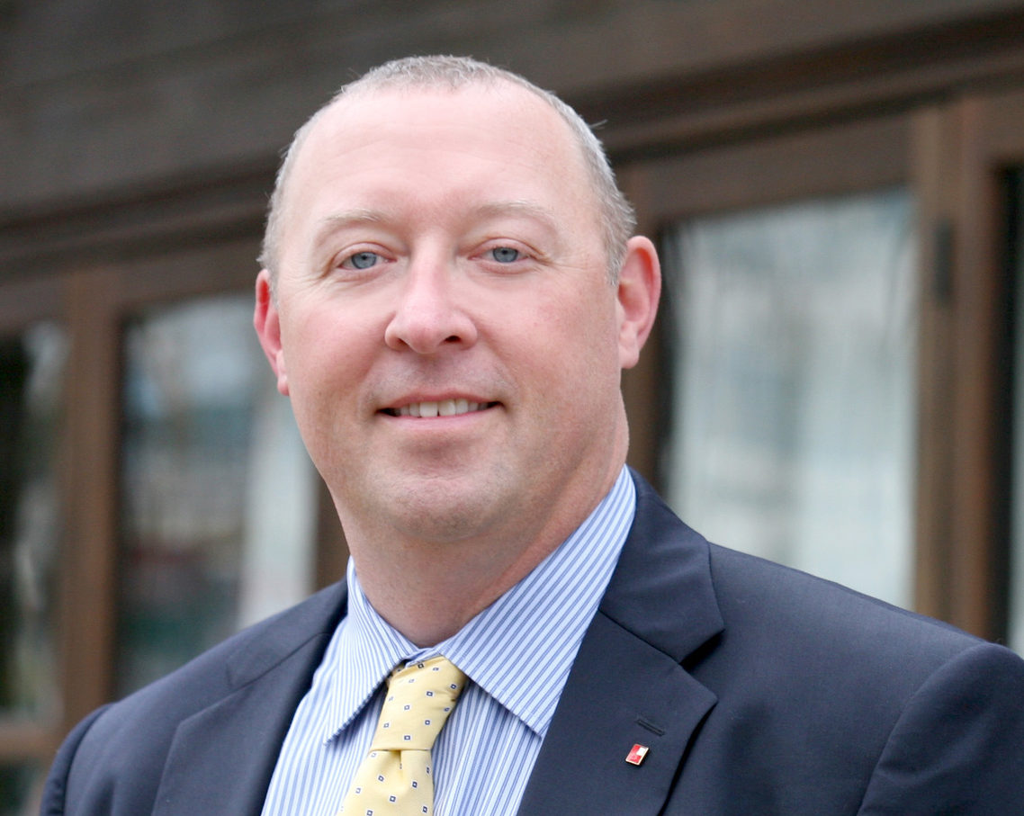 smiling man in a dark suit and yellow tie in front of glass doors