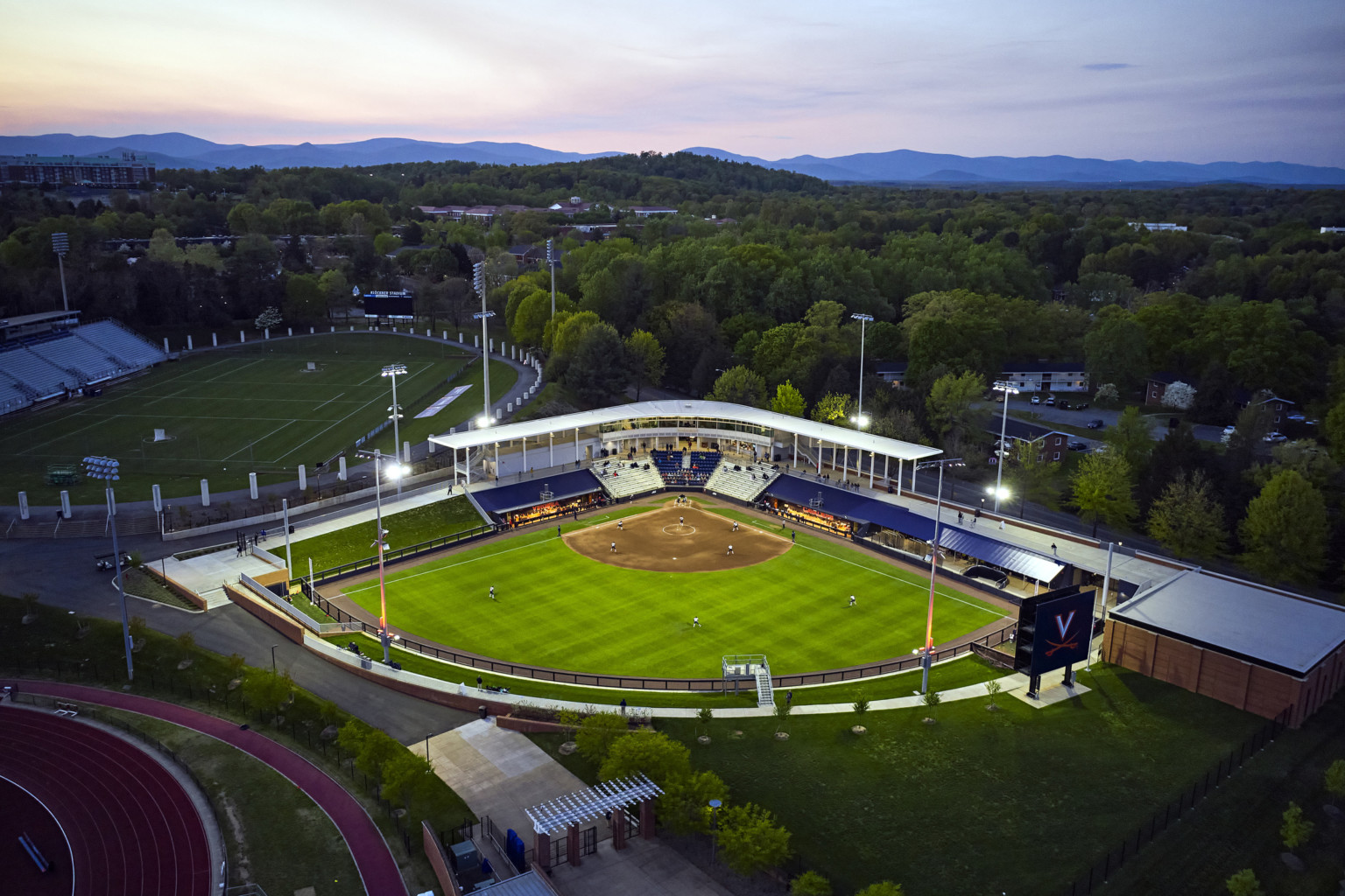 Aerial view of softball stadium at dusk lit up but lights and surrounded by lush trees Aerial view of softball stadium at dusk lit up but lights and surrounded by lush trees