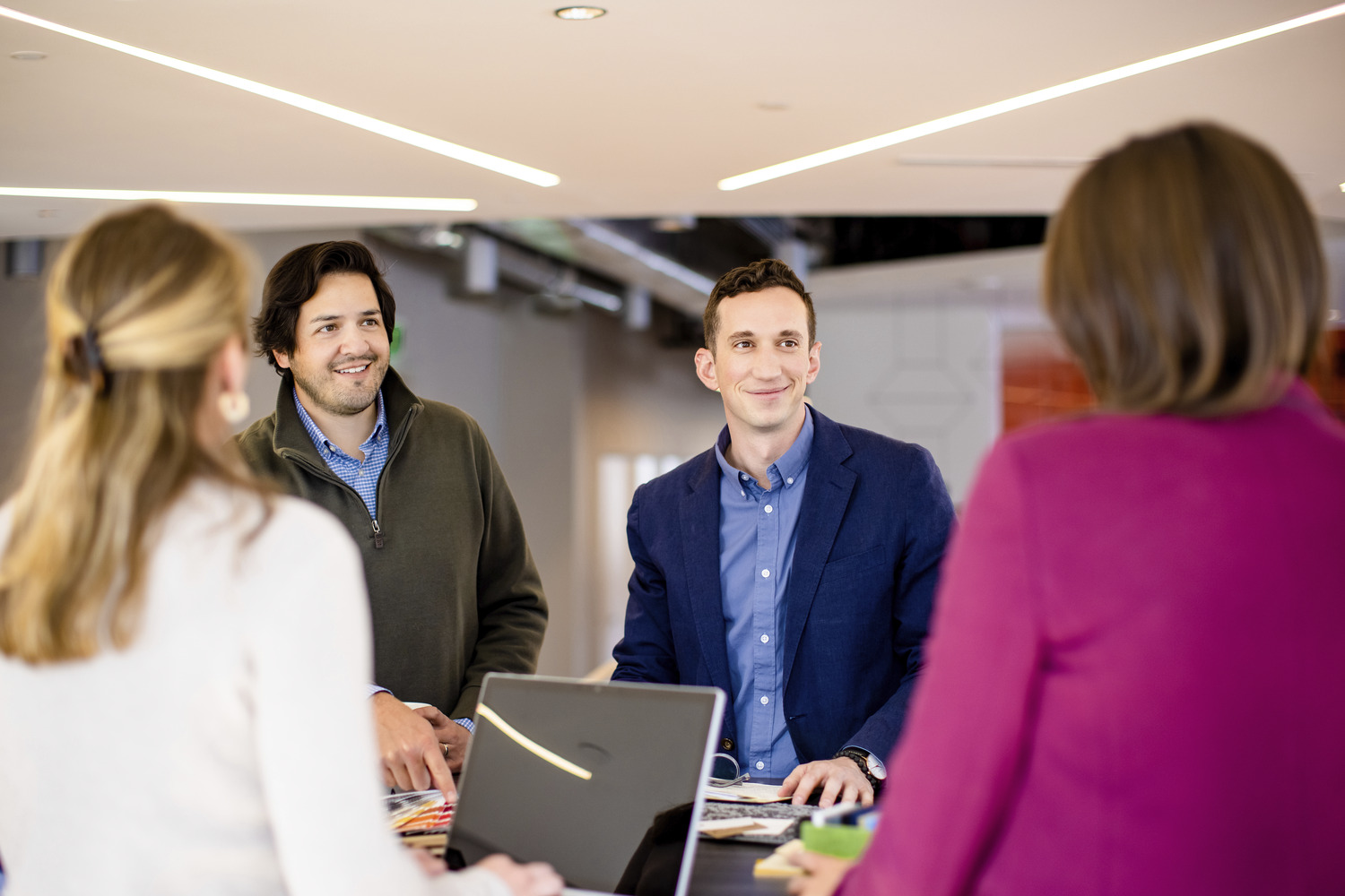 a group of smiling young professionals collaborating around a standing height table under a ceiling with diagonal lighting